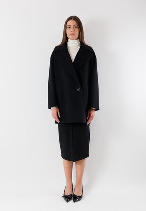 Cappotto oversize in misto lana nero con rever pronunciati, chiusura a singolo bottone, abbinato a una gonna nera al ginocchio e décolleté nere a punta.