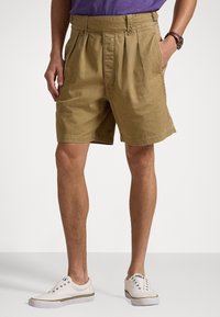 Man i beige veckade shorts, vita slip-on sneakers och en lila skjort, står med ena handen i fickan mot en neutral bakgrund.