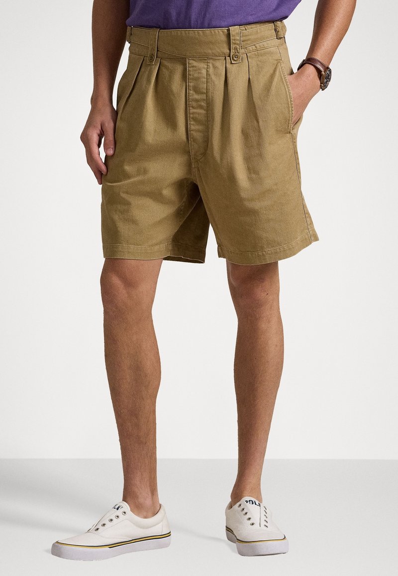 Man i beige veckade shorts, vita slip-on sneakers och en lila skjort, står med ena handen i fickan mot en neutral bakgrund.