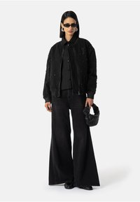 Černá bomber jacket s cvočkovými ozdobami, černé tričko, široké džíny a strukturovaná černá tkaná kabelka. Nosené tmavé sluneční brýle.