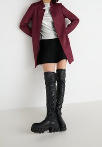Burgunderfarbenes langes Kleid über einem silbernen Pailletten-Oberteil, kombiniert mit einem schwarzen schwingenden Rock und kniehohen schwarzen Lederstiefeln mit dicken Sohlen.