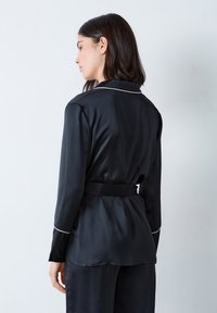 Czarna satynowa bluzka od piżamy z długimi rękawami, zdobiona białą lamówką przy kołnierzu i związanym paskiem z tyłu, co podkreśla sylwetkę. Gładka tekstura.