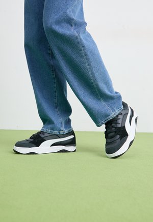 Zapatillas en negro y gris con un acento blanco; cuentan con una parte superior de malla texturizada, cuello acolchado y suela gruesa blanca. Combinadas con vaqueros azules.