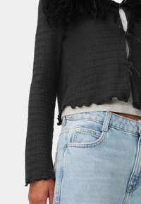 Zwarte textuur geknipte cardigan met ruffles aan de onderkant en strikjes aan de voorkant, gecombineerd met lichtblauwe denim jeans.