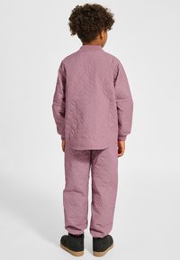 Dziecko o kręconych włosach nosi pikowaną jasnofioletową kurtkę i dopasowane spodnie, stoi tyłem na białym tle, ma czarne buty.