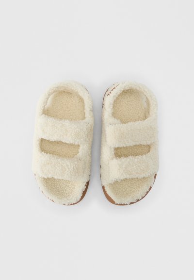 Sandales à enfiler couleur crème avec une texture douce et duveteuse. Deux brides ajustables à Velcro et une semelle plate de couleur beige. Convient pour une utilisation à l'intérieur ou pour un port décontracté.