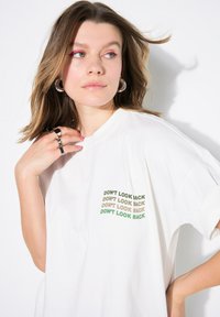 Camiseta blanca de gran tamaño con el texto "NO MIRES ATRÁS" en verde, marrón y negro en el lado izquierdo del pecho. Textura de algodón suave.