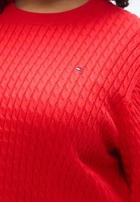 Κόκκινο πουλόβερ με πλέξη καλωδίου, φτιαγμένο από απαλό ύφασμα, με ριμπ λαιμόκοψη και μικρή λεπτομέρεια λογοτύπου στο στήθος.