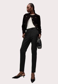 Zwarte getextureerde jas met gouden knopen, wit topje, getailleerde zwarte broek en zwarte pumps met puntige neus. Houdt een kleine zwarte handtas vast.