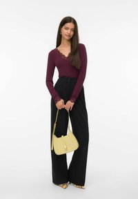 Femme avec de longs cheveux portant un haut bordeaux bordé de dentelle, un pantalon noir taille haute à jambes larges, des chaussures à imprimé serpent, tenant un sac à main jaune.