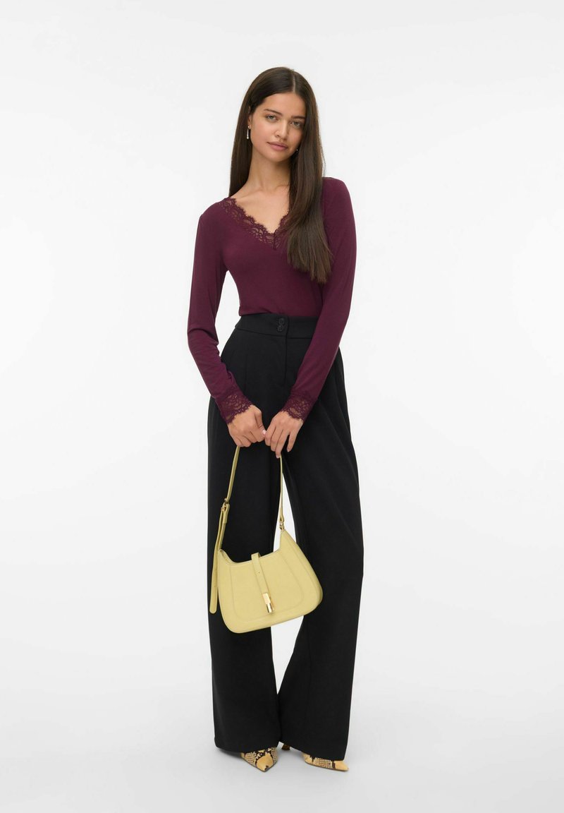 Femme avec de longs cheveux portant un haut bordeaux bordé de dentelle, un pantalon noir taille haute à jambes larges, des chaussures à imprimé serpent, tenant un sac à main jaune.