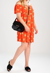 Schulterfreies oranges Kleid mit gelbem Blumenmuster, kombiniert mit einer schwarzen Bag aus Lackleder und schwarzen Rutschsandalen mit Rüschendetail.