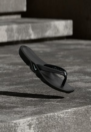 Tong noir Nike flottant au-dessus d'un sol en béton texturé avec des marches en arrière-plan, vue de côté et de dessus.