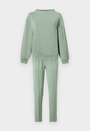 Měkká zelená mikina s dlouhými balónovými rukávy a odpovídající přiléhavé kalhoty s předními švy na bílém pozadí.