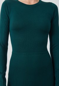 Rochie cu mâneci lungi, de culoare teal profund, cu o textură canelată pe partea superioară și o fustă pliată, având un decolteu rotund.