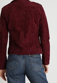 Bordeaux suède jasje met gestructureerde naden, schouderepauletten en een getailleerd ontwerp. Draag het over hoge taille blauwe jeans met achterzakken.