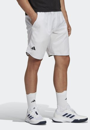 Personne portant un short de sport blanc avec un logo noir, des chaussettes blanches montantes et des baskets blanches à rayures noires, debout les mains dans les poches.