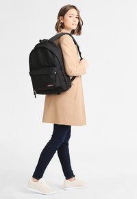 Schwarzer Stoff-Rucksack mit verstellbaren Trägern, vorderer Reißverschlusstasche und Logo. Wird von einer Person in einem beigen Mantel und dunklen Jeans getragen.