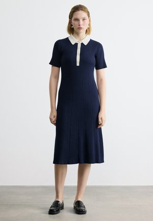 Γυναίκα όρθια, φοράει ένα μπλε σκούρο φόρεμα με ριπ ύφασμα, κοντά μανίκια και λευκό γιακά, σε συνδυασμό με μαύρα μοκασίνια σε γκρι δάπεδο.