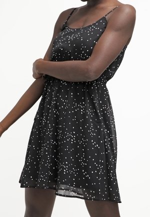 Femme portant une robe noire sans manches avec un motif blanc tacheté, dotée de fines bretelles et d'une taille cintrée.