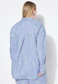 Camicia a righe blu e bianca con ricami floreali verdi, colletto con bottoni e maniche lunghe, caratterizzata da una vestibilità rilassata e un dettaglio arricciato sul retro.