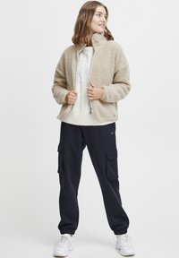 Beige fleecejakke med høy krage, glidelås og ribbede mansjetter, kombinert med en hvit kabelstrikket genser og marineblå cargo-bukser.
