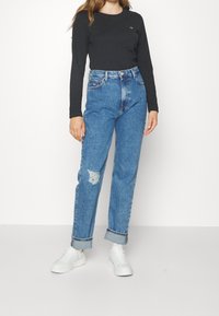 Zwarte langärmmuts, hooggesneden blauwe jeans met beschadigde plekken, opgerolde zomen en witte sneakers. Effen achtergrond.