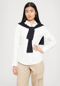 Giovane donna che indossa una camicia bianca con bottoni e pantaloni cargo beige, con un maglione scuro appoggiato sulle spalle, in posa con una mano sul fianco.
