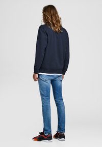 Sweatshirt marine avec des poignets côtelés, jeans bleu clair et chaussures de sport noires/rouges. Le modèle est vu de dos contre un fond uni.