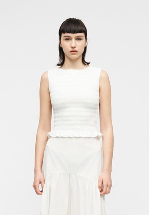 Γυναίκα με κοντά σκούρα μαλλιά που στέκεται, φορώντας λευκό αμάνικο τοπ με σφηκοφωλιά στο σώμα και βολάν στο τελείωμα, σε συνδυασμό με λευκή φούστα.