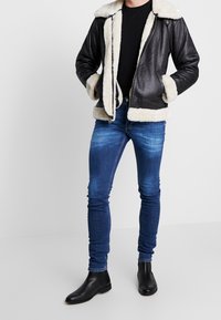 Zwarte leren jas met een crèmekleurige schapenvachtkraag en -manchetten, gecombineerd met blauwe slim-fit jeans en zwarte enkellaarzen, staand tegen een neutrale achtergrond.