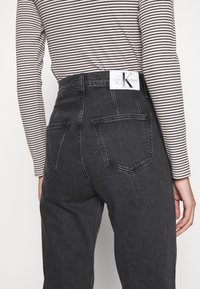 Tmavě šedé džíny z denimu s vysokým pasem, s dvěma zadními kapsami. Na pásu je viditelný bílý štítek s logem.