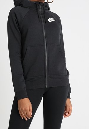 Kobieta w czarnej bluzie Nike na suwak z przednimi kieszeniami i czarnych legginsach, stojąca na tle jednolitego białego tła.