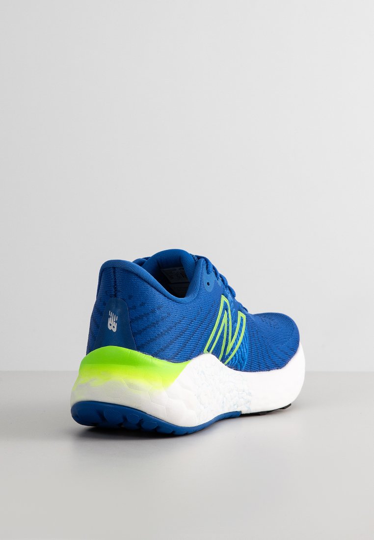Zapatilla de correr New Balance azul con suela verde neón y blanca, vista desde el lado trasero izquierdo sobre una superficie gris.