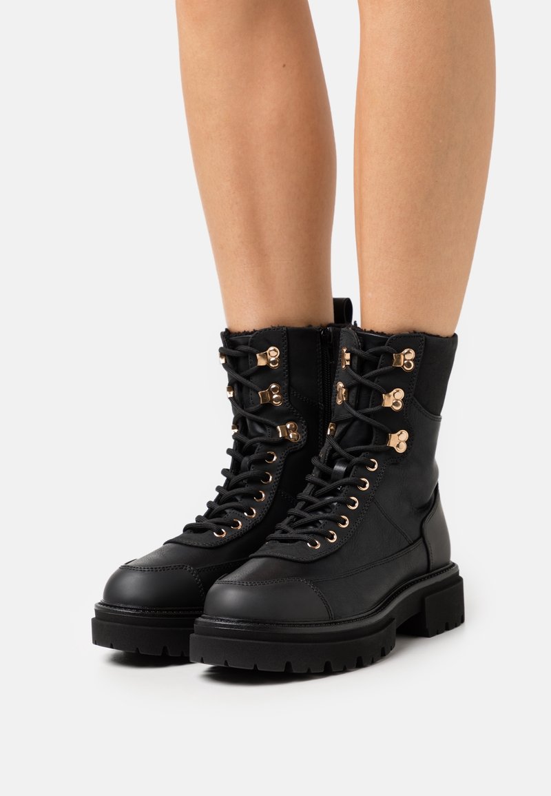 Botas de cuero negro hasta el tobillo con punta redonda, acentos en dorado, cordones en la parte delantera y un cuello texturizado, que cuentan con una suela robusta.