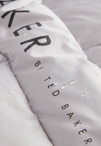 Weißes Steppjacken-Material mit sichtbaren Wassertropfen. Auffälliger schwarzer Text mit der Aufschrift "TED BAKER" und "by TED BAKER." Leichtes Material mit strukturierter Oberfläche.