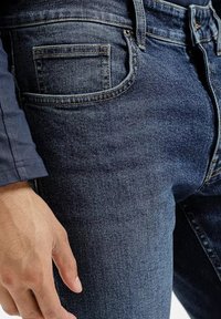 Ciemnoniebieskie dżinsy z widokiem z bliska na kieszeń, z metalowymi elementami w kolorze srebrnym i widocznym przeszyciem. Tkanina ma gładką fakturę.