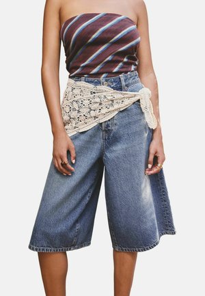 Person iført vidde-ben denimshorts med et cremefarvet blonderem bundet ved taljen og en skulderløs brunstribet top.