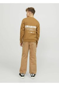 Sudadera marrón con gráfico en blanco "COLLECTIVE" en la parte posterior, combinada con pantalones de pierna ancha color tostado y zapatillas negras con acentos en blanco.
