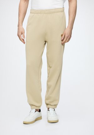 Man wearing beige elastic-waist jogger pants and white sneakers, standing against a plain white background.