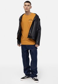 Veste en cuir noir sur un t-shirt orange avec impression "GRACIOUS", jeans en denim bleu avec patchwork, et baskets noires avec semelles blanches.
