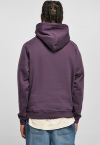 Paarse hoodie met een grote capuchon, ribbelmanchetten en onderrand, textiele stof en een eenvoudig ontwerp, gedragen over een wit shirt en jeans.