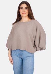 Helles Taupe, lässiges Oberteil aus strukturiertem Stoff mit weiten Ärmeln und Spitzenakzenten am Saum, kombiniert mit blauen Jeans.