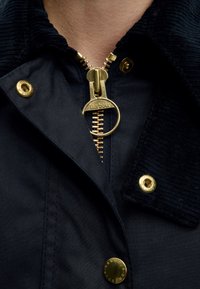 Gros plan d'une veste sombre avec une fermeture éclair dorée et des boutons, avec un grand tirette circulaire gravée du nom de la marque Barbour.