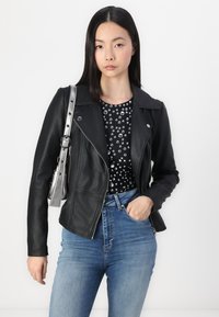 Chaqueta de motociclista de cuero negro con cremalleras, solapas en pico y diseño estructurado, combinada con una blusa de lunares en blanco y negro y jeans de cintura alta.