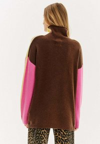 Hnedý sveter s vysokým golierom a rukávmi v ružovej a žltej farbe. Mäkkučká textúra, voľný strih. Nosené s nohavicami v leopardom vzore.