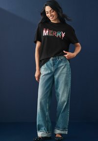 Čierne tričko s nápisom "MERRY" v farebných písmenách, skombinované s svetlomodrými oversized džínsami a čiernymi balerínkami. Tmavomodré pozadie.