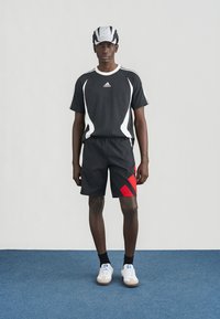 Sportoutfit met een zwart t-shirt met witte accenten en drie strepen, gecombineerd met zwarte shorts met een rood diagonale patroon.