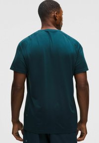 Zöld aktív póló texturált anyagból, rövid ujjakkal és kerek nyakkivágással; a szegélye közelében finom márkázás található.