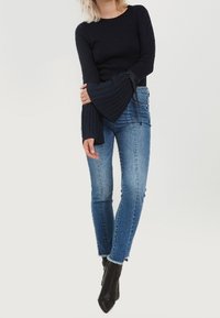 Une femme portant un haut noir à manches longues avec des poignets évasés, un jean bleu ajusté et des bottines noires, debout devant un fond uni.